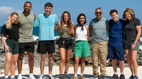 BBC The eight celebrity contestants standing in a row with their arms around each other in front or rocks and the sea