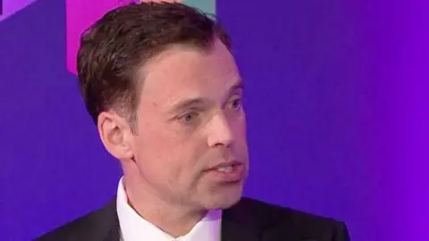 Ken Skates speaking in the debate in Wrexham, wearing a dark suit and red and white striped tie