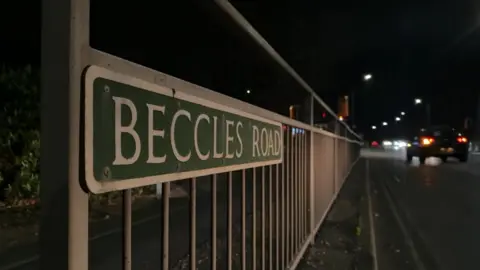 Andrew Turner/BBC A road sign stating Beccles Road, lit by car headlights. The sign shows white text and border on a green background and the sign is affixed to metal railings. Car tail lamps and headlights can be seen to the right of the image.