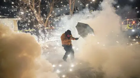 A protester on a snowy street uses an umbrella to shield themself from a cloud of tear gas and sparks in Minneapolis