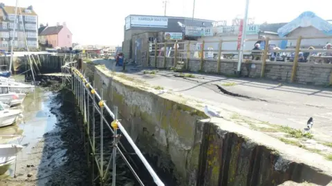 The view along the crumbling harbour wall which is lined with scaffolding. The tide is out and several small boats are resting on the mud below. The tarmac path on top of the wall is wobbly with cracks along its length. The side of the concrete wall is also visibly crumbling. On the land side of the path is a low brick wall. Beyond the wall are food outlets and people sitting at picnic benches.
