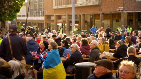 A photo of Coventry's Broadgate square with people sat watching the event on picnic benches. In the foreground, a woman is wearing the blue and yellow flag of Ukraine