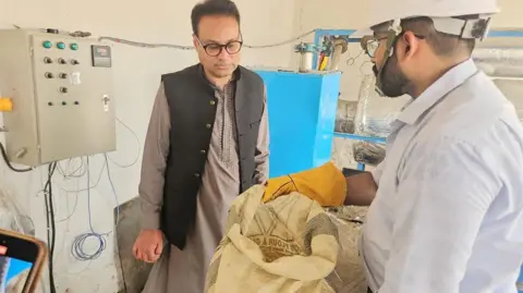 Northumbria University Dr Jibran Khaliq at a demonstration event held in Saleh Pat, Pakistan. In a laboratory setting, a man dressed in a white shirt and PPE holds a white sack while Dr Khaliq looks at its contents.