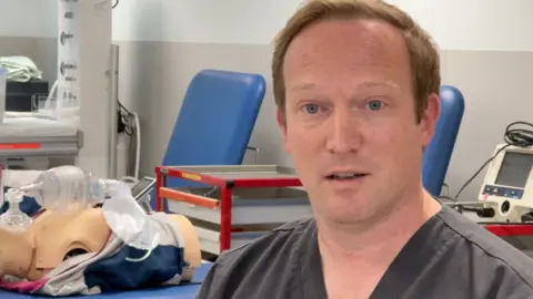 Andrew Blackmore looks serious as he sits in a hospital room, surrounded by medical equipment. He has short red hair and wears a gray apron. 