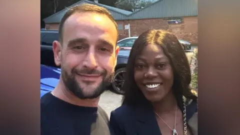 NIHR Richard Oldale, 42, from Bradford with his fiancée Chrystelle - they are stood in front of a building and some parked cars, smiling 
