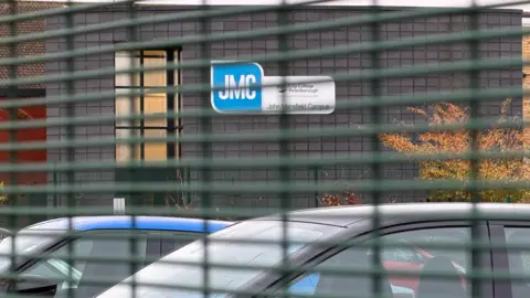 A general view of the John Mansfield Centre through a wire fence. There are cars in a car park in the foreground and a building with a "JMC" sign in the background.