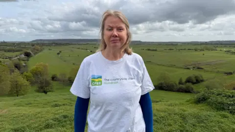 A woman in a grey t-shirt with the CPRE logo on over a blue shirt is looking at the camera with a neutral expression. Behind her a green fields rolling into the background, with a grey sky above.