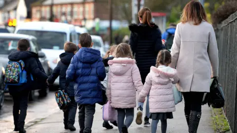 Children walking to school with their parents. They are facing away from the camera. There are three boys on the left of the picture wearing blue overcoats, while two girls on the right of the picture are wearing pink hooded coats.