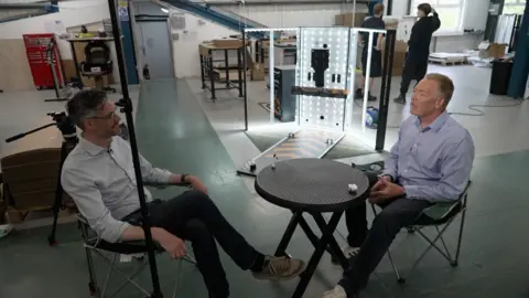 Andy Meeson/BBC Two people sitting inside, around a round plastic table inside an industrial warehouse. Andrew Moss is sat on the right and appears to be mid-sentence. He is wearing a blue shirt and dark trousers. Ben Schofield, a BBC reporter, is sitting on the left, facing Moss. He is wearing a light shirt and dark trousers. He is sitting with his legs crossed and is partially obscured by the pole from a lighting stand. Behind Schofield is a camera tripod, with no camera attached. In the middle of the frame and behind them both is a piece of equipment festooned with lights shining out of it. Two workers in dark clothing can be seen behind the equipment. 