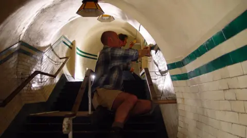 Mike Tucker Two men are sat in a miniature version of a London tube tunnel painting the walls the correct colour.