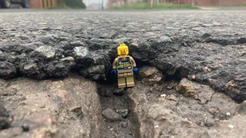 A Lego minifgure stands in a damaged road surface.