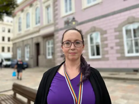 Deputy Louise Doublet in a purple top and glasses stands in front of a pink granite building, Jersey's States Assembly.