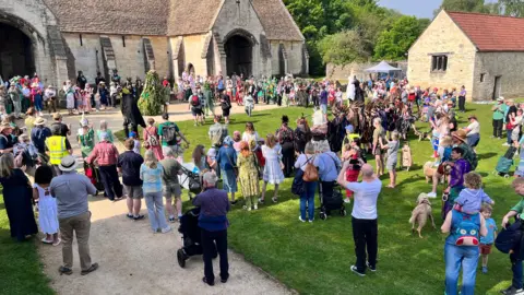 Large group of people standing outside on grass in front of a historic church-like building. They are all watching a large figure in green in the middle of the circle they have made.