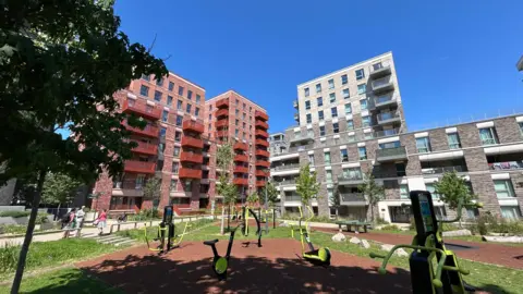 A wide angle shot of three housing blocks - the two on the left are made of red brick and the one on the right is light grey/brown bricks. In the foreground is a play area.