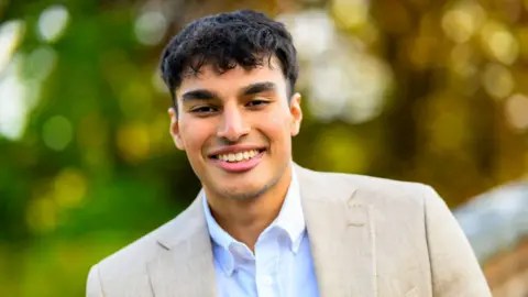 Faraaz Noor smiling at the camera. He has wavy black hair and is wearing a white shirt and a beige suit jacket.