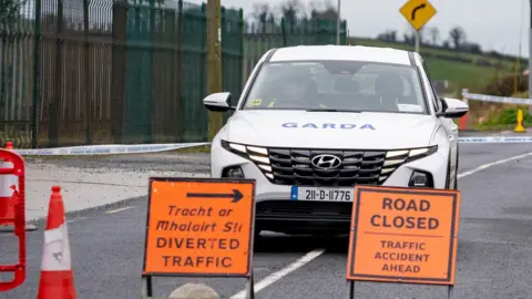 PA Media A garda car is parked on a road with a garda officer in the driver's seat. An orange road sign sign says road closed, traffic accident ahead in English. Another sign in Irish and English says diverted traffic. An orange cone is sitting on the road and police tape is up. 
