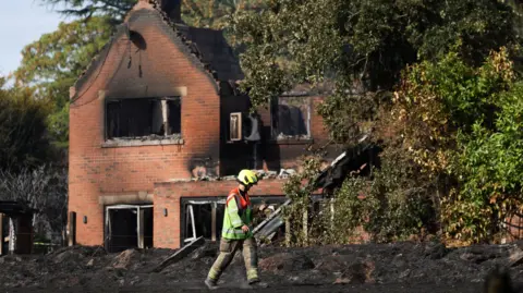 Reuters A firefighter in brown trousers and an orange and yellow hi-vis jacket with a yellow helmet is walking in front of a house that has been destroyed by fire. It has black marks on its exterior and the windows have blown out