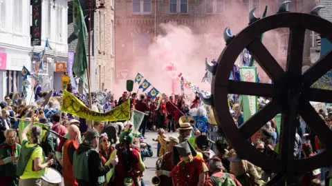 The Hellfire Kernow event in Redruth. The street is filled with people who are wearing costumes and holding banners. There is a wooden wheel in the foreground which has spikes around its and there are clouds of red smoke.