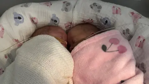 Two premature babies lie asleep, face-to-face. One is swaddled in a white, knitted blanket, the other is swaddled in a pink, fleece blanket. Both babies have breathing tubes taped to their faces.