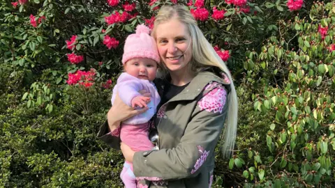 Alan Johnstone Anneka holding her baby daughter Sienna, who is wearing a pink outfit and pink bobble-hat. The mother and daughter are smiling and standing in front of green trees with bright pink flowers.