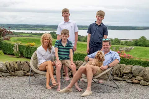 IWC Media A group of five people sit and stand together outdoors on gravel beside a low stone wall, accompanied by a small dog resting on a seated person’s lap.
Behind them stretches a lush green landscape with rolling fields and a calm body of water under a cloudy sky.
Casual seating, light clothing, and garden flowers create a relaxed, scenic setting in the countryside.
