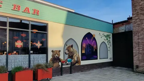 A mural of two bears enjoying a hot chocolate has been painted on the side of a cafe bar in Kenilworth. In another panel Kenilworth Castle has been painted with fireworks seen going off in the background. Ivy has also been painted as part of the mural. 