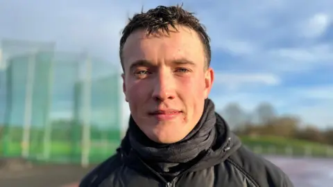 A young man with short dark hair, wearing a black jacket and a black scarf, looks at the camera.