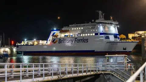 Brittany Ferries A picture of a Britttany Ferries Ferry parked at St Peter Port harbour at night, it is white and blue, and behind it you can see grey Castle Cornet lit up by yellow light. 