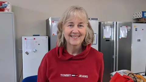 Foodbank trustee Susan Hornby with blond collar-length hair stands in front of the freezers wearing a red hooded top