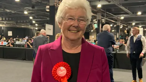 BBC A woman with short white hair wears a pink jacket and a black top and wears a red Labour Party rosette. Behind her are tables and people sat or standing around.