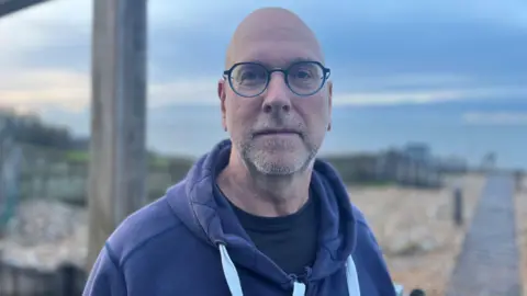 BBC / George Carden A bald man wearing glasses and a blue hooded top and t-shirt stands on a beach