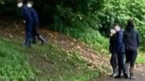 Five young children at a grassy slope in a park near trees. Three stand on a path and two are on the slope. All have their faces blurred.