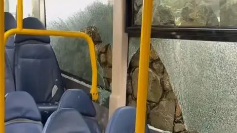 A hole can be seen in a window and glass surrounding it has shattered but not broken. The image was shot inside a bus and blue seats are in shot, with a shard of glass on one of them.