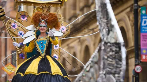 Mark Waugh A woman dressed as a giant queen bee as part of a parade. Her dress is yellow and black with giant wings and wearing a crown. She is in a city centre street. She is smiling.   