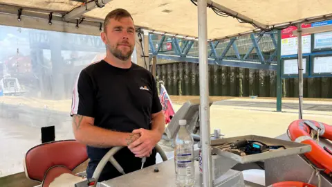 BBC Ben Ellis stands behind the steering wheel of the King's Lynn Ferry. He is wearing a black top and has tattoos on his forearm.