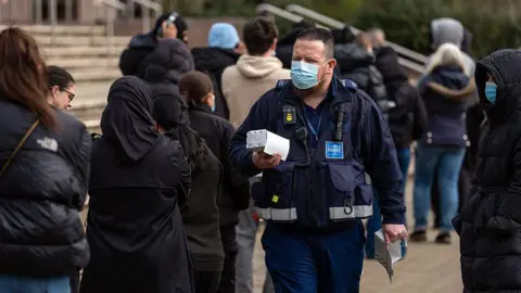 Getty Images A man in a dark navy uniform with a high-visibility trim and a University of Kent lanyard walks past a queue of people, wearing a blue surgical face mask. He holds papers and a small box in his hands, with a body-worn camera and radio visible on his chest. Behind him, a line of young people in dark winter coats stand with their backs to the camera, some also wearing face masks, waiting outside what appears to be a campus building.