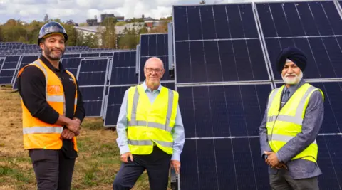 Three men are in the foreground looking at the camera, with Rennalls on the left wearing an orange hi-vis jacket and the other two wearing a yellow hi-vis jacket. They are standing in front of a line of blue panels going from the right back to the far left, with trees in the background.