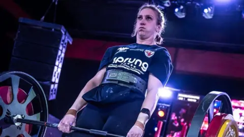 Jade Skill lifts a barbell with various weights on each end. She has dark hair that has been plaited back and wears a black T-shirt with chalk over it. Her cheeks are puffed out with the effort of lifting the weight.