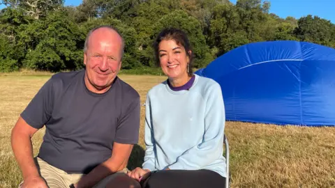 Jo is sat next to her husband, Clive, on a filed with a hot air balloon lying next to them on the ground. 