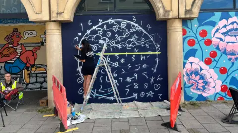 An artist paints a navy blue space with various patterns and symbols. To her right is a mural with flowers and cherries on and to her left is another mural depicting a woman. She is stood on a ladder.