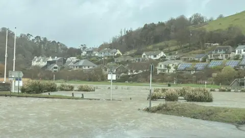 Josh A car park in Cornwall in Seaton is flooded, the floor is water and there are houses sat behind it, some with blue solar panels.