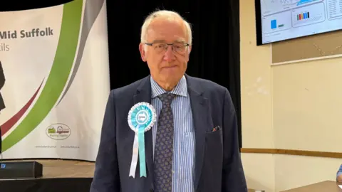 Louise Parker/BBC A head and shoulders image of a man wearing a Reform rosette on a navy blazer over a striped blue and white shirt and patterned blue tie. He is looking into the camera. 
