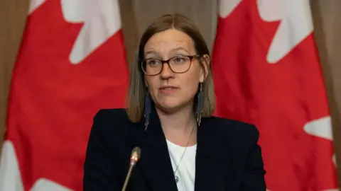 Bloomberg via Getty Images Karina Gould in a white shirt and dark suit coat sits before a microphone and Canada flags during a press conference in Ottawa, Canada, on April 19, 2023