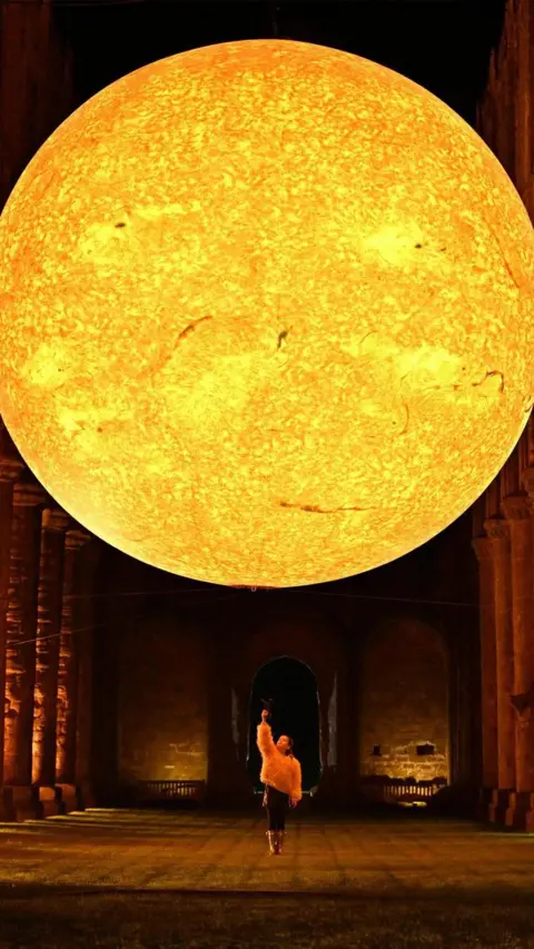 National Trust Images, Anthony Chappell-Ross A portrait photo of a large sun display suspended above pillars in a church-like building. Below the sun depiction is a girl looking up at it with her arm pointing up to it. She looks very small compared to the sun display.