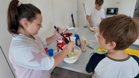 Three children a young girl at the front left a young boy at the front right and a young boy in the distance. They are stood around a table with mixing bowls and baking ingredients on the table.