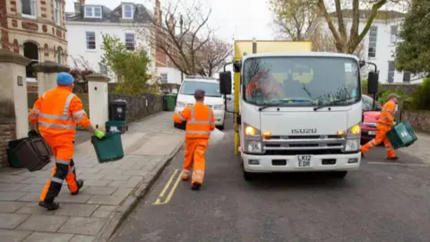 Refuse collectors wearing hi-vis orange suits on a residential road putting recycling into a refuse truck.