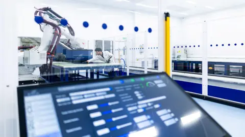 ICOMAT An engineering facility with an interactive screen in the foreground and an engineer preparing a robotic arm in the background.