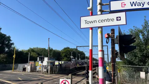 Railway tracks with overhead cable stretch off into the distance. In the foreground, they are intersected by a level crossing. There is also a red and white striped pole with a sign attached which reads "town centre" alongside an arrow pointing right. A sign above it reads "way out". 