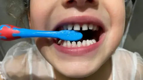A close-up of a child's mouth with a blue toothbrush in between her teeth