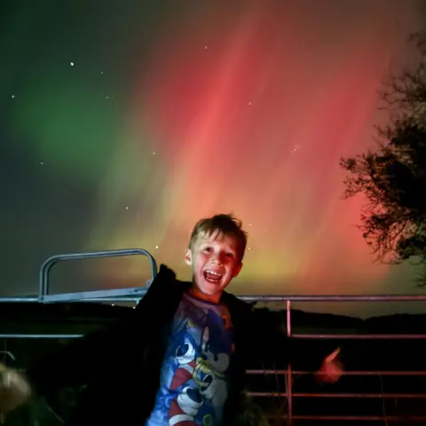 Emma Paliczka Youngster Aaron Paliczka has a big smile on his face as the aurora appears in green and pink behind him.